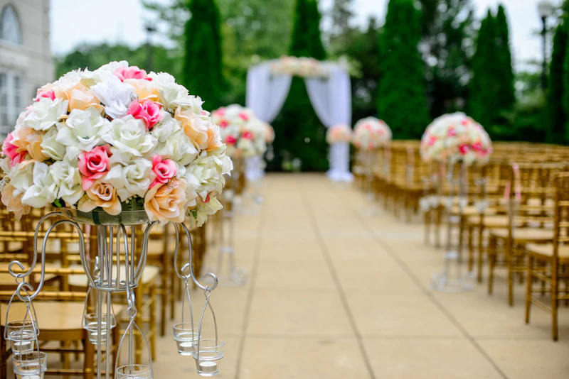 Outdoor ceremony aisle lined with tall rose-and-greenery arrangements and gold chairs, a draped altar beyond