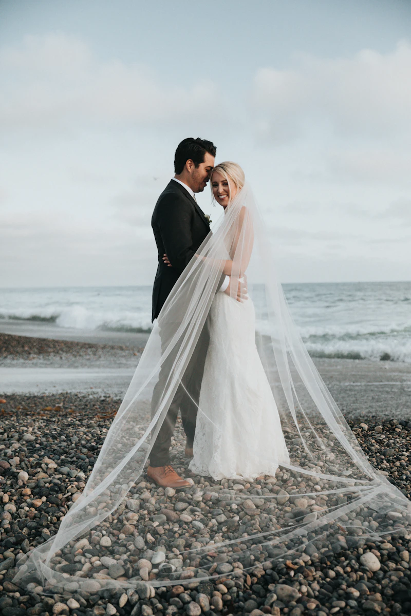 Couple on a pebble beach at the water's edge, the bride's long veil sweeping across the stones