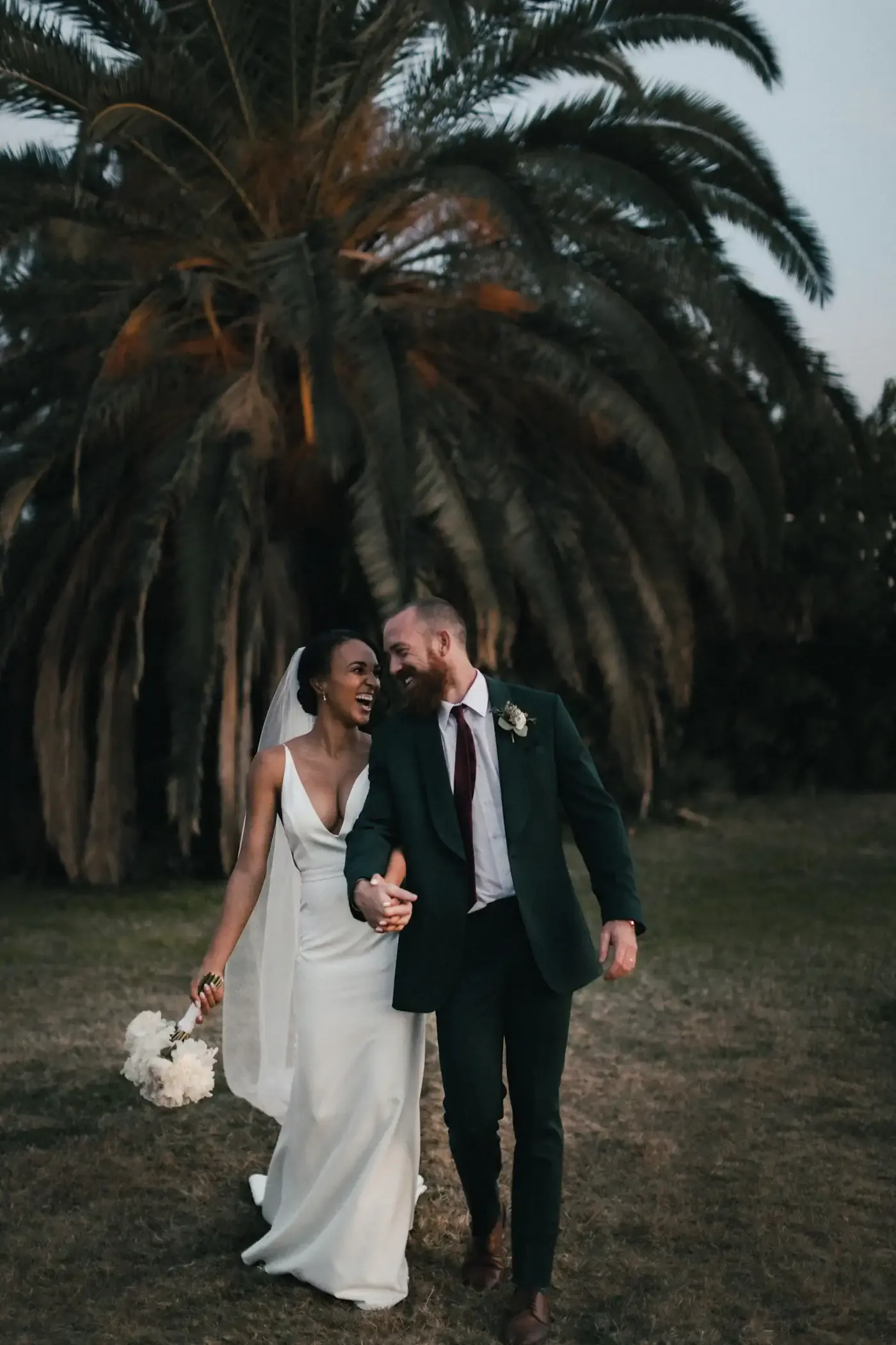 Bride and groom walking hand in hand and laughing beneath a palm tree at dusk